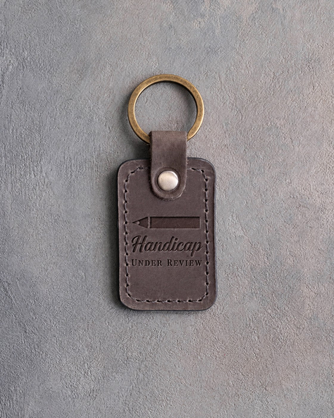 Country gray leather keychain debossed with a scorecard pencil above Handicap UNDER REVIEW, on gray plaster