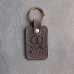 Country gray leather keychain debossed with two interlocked wedding rings above the words Has the RINGS, on gray concrete