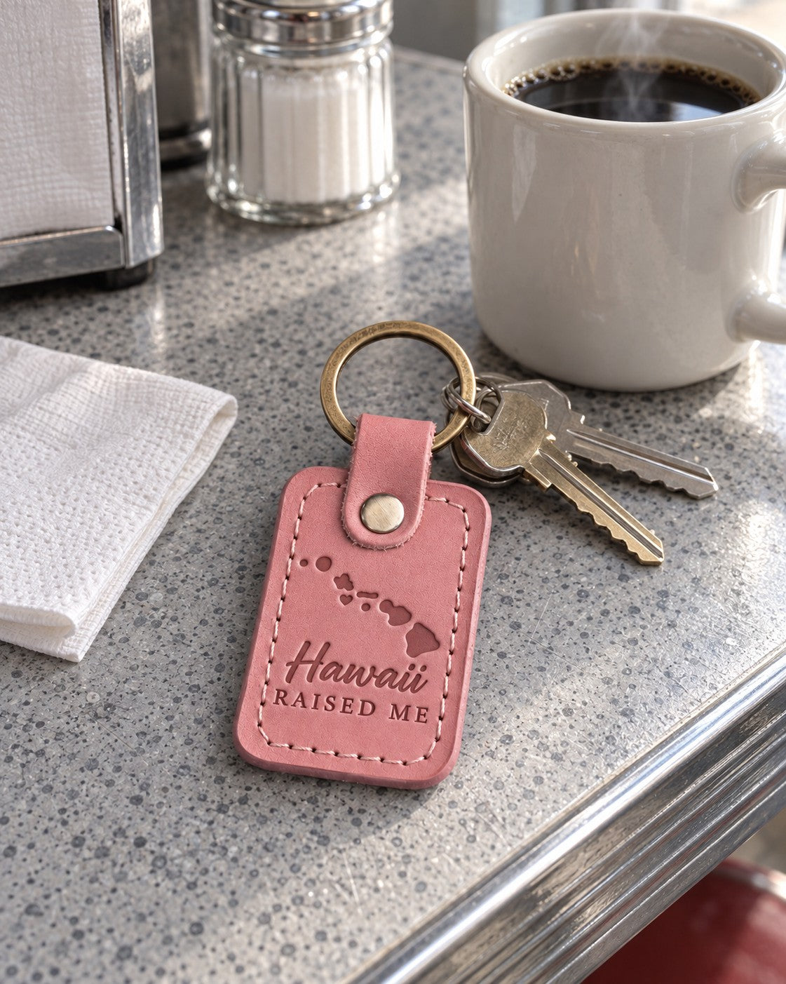 Pink Hawaii Raised Me keychain on apartment keys beside a plumeria lei on a mainland kitchen counter