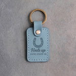Turquoise leather keychain debossed with a heels-up horseshoe above Heels up and LUCK STAYS IN, on gray plaster