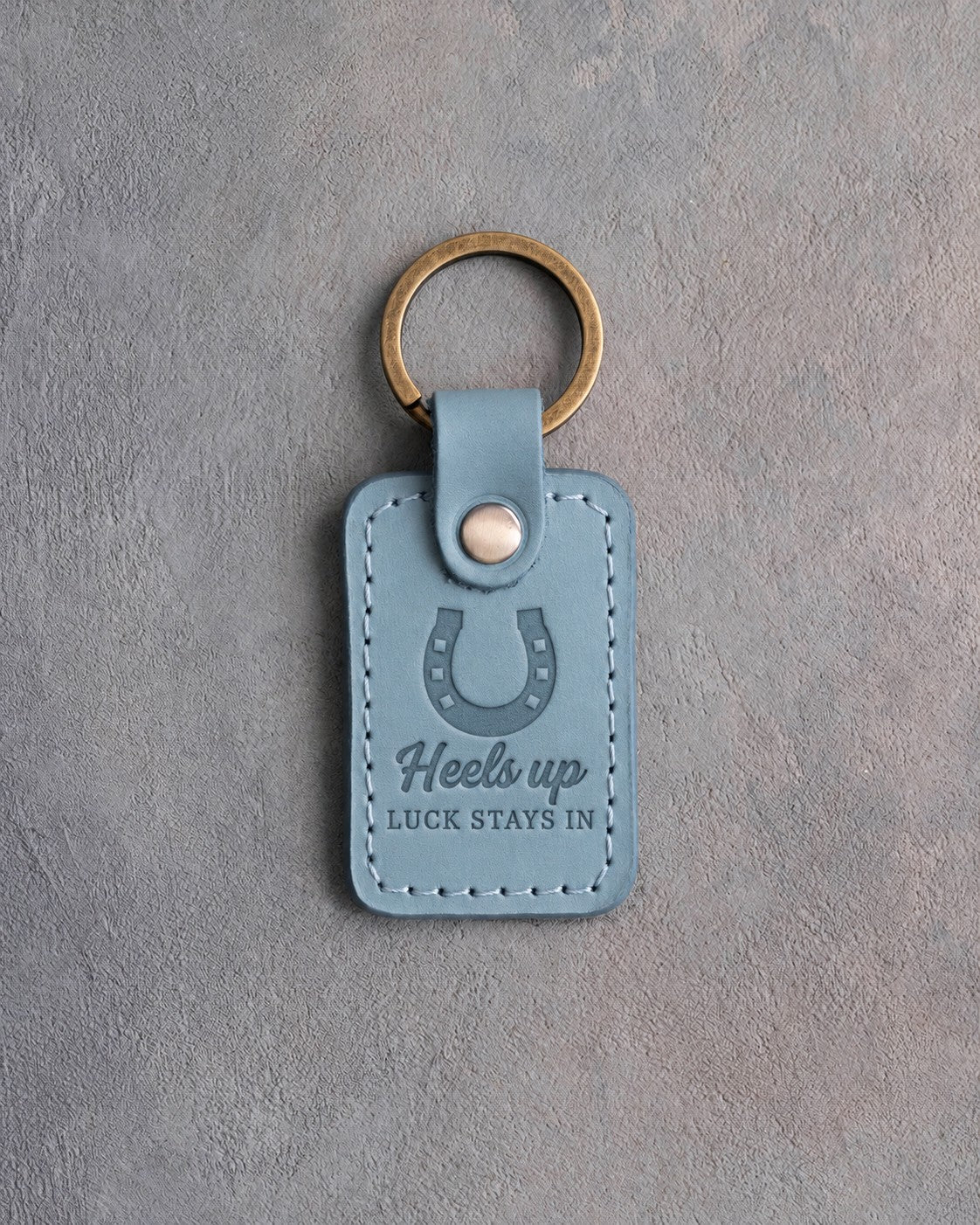 Turquoise leather keychain debossed with a heels-up horseshoe above Heels up and LUCK STAYS IN, on gray plaster