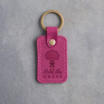 Fuchsia leather keychain debossed with a hand-tied bouquet above the words Held the DRESS, on gray concrete
