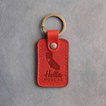 Poppy red leather keychain debossed with a California silhouette and a heart at San Francisco above Hella and NORCAL, on gray concrete