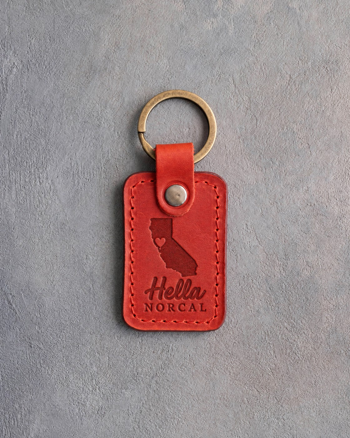 Poppy red leather keychain debossed with a California silhouette and a heart at San Francisco above Hella and NORCAL, on gray concrete