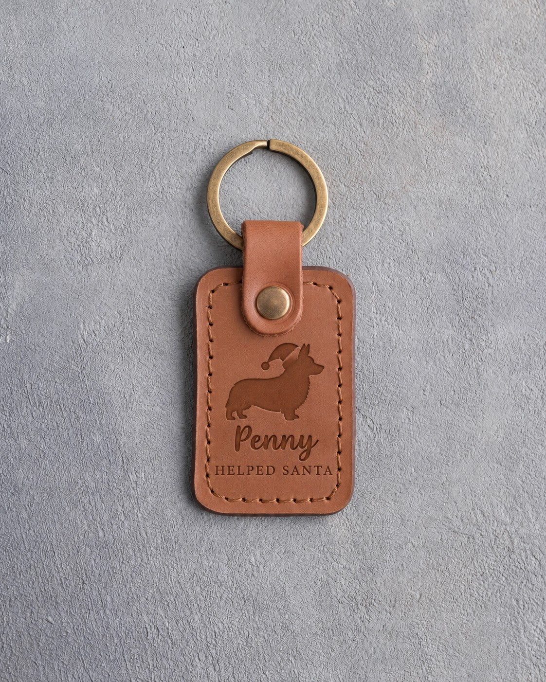 Caramel leather keychain debossed with a corgi in a Santa hat above Penny and HELPED SANTA, on gray concrete