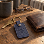 Her HUSBAND keychain in navy leather on an oak kitchen table beside a worn bifold wallet and a stoneware mug