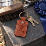Tan Clover The Highland keychain on a painted show box lid in a livestock barn alley beside a show stick and a blue rosette