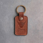 Arizona tan leather keychain debossed with a shaggy highland cow above the name Clover and THE HIGHLAND, on gray concrete