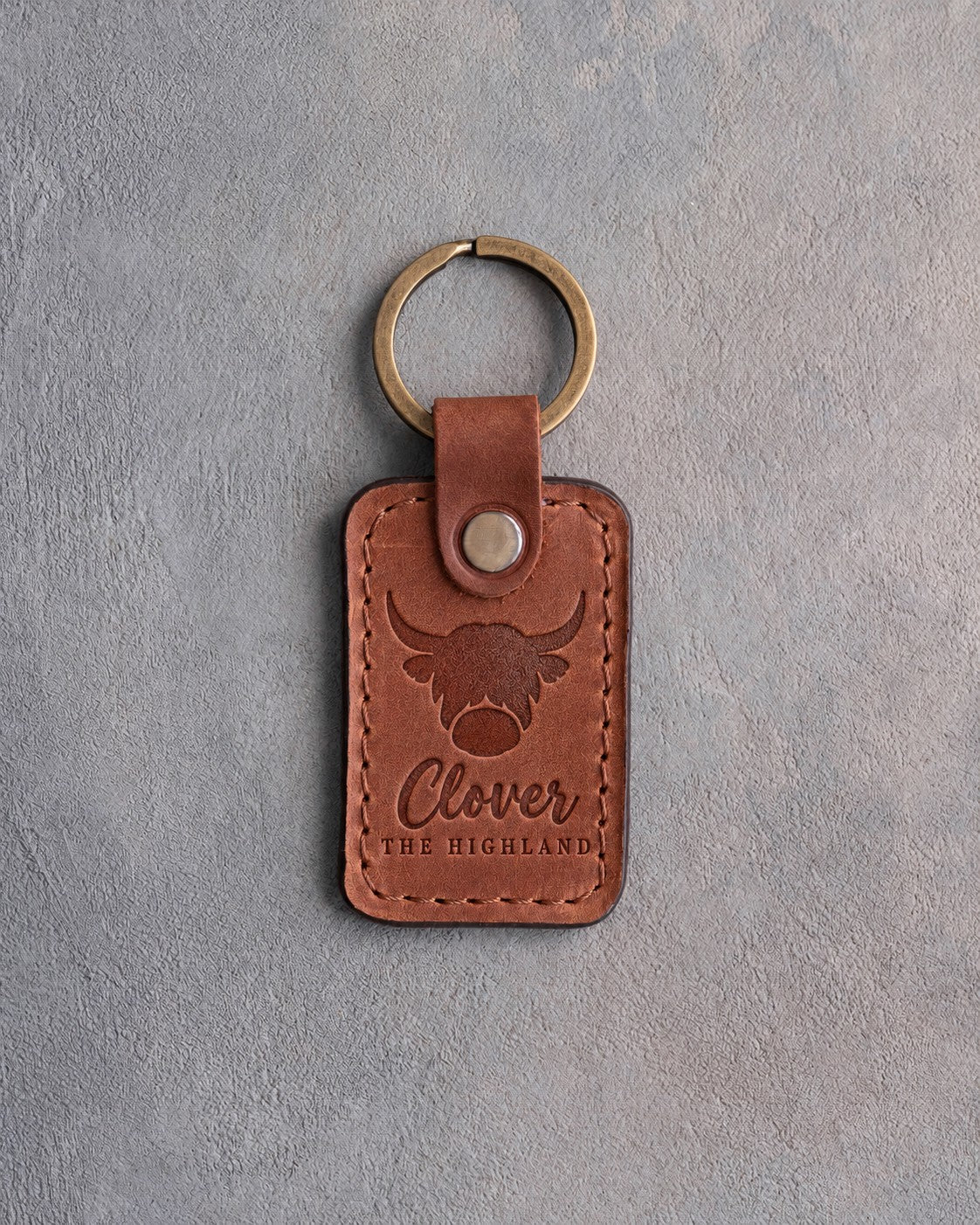 Arizona tan leather keychain debossed with a shaggy highland cow above the name Clover and THE HIGHLAND, on gray concrete