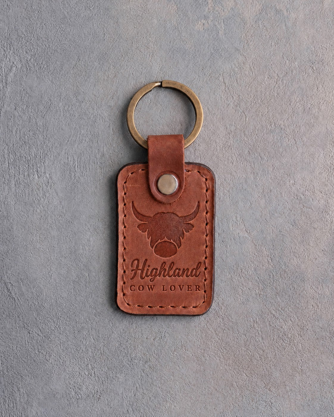 Arizona tan leather keychain debossed with a shaggy highland cow above the words Highland and COW LOVER, on gray concrete