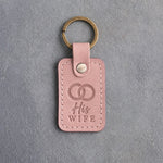 Blush pink leather keychain debossed with two interlocked wedding rings above the words His WIFE, on gray concrete