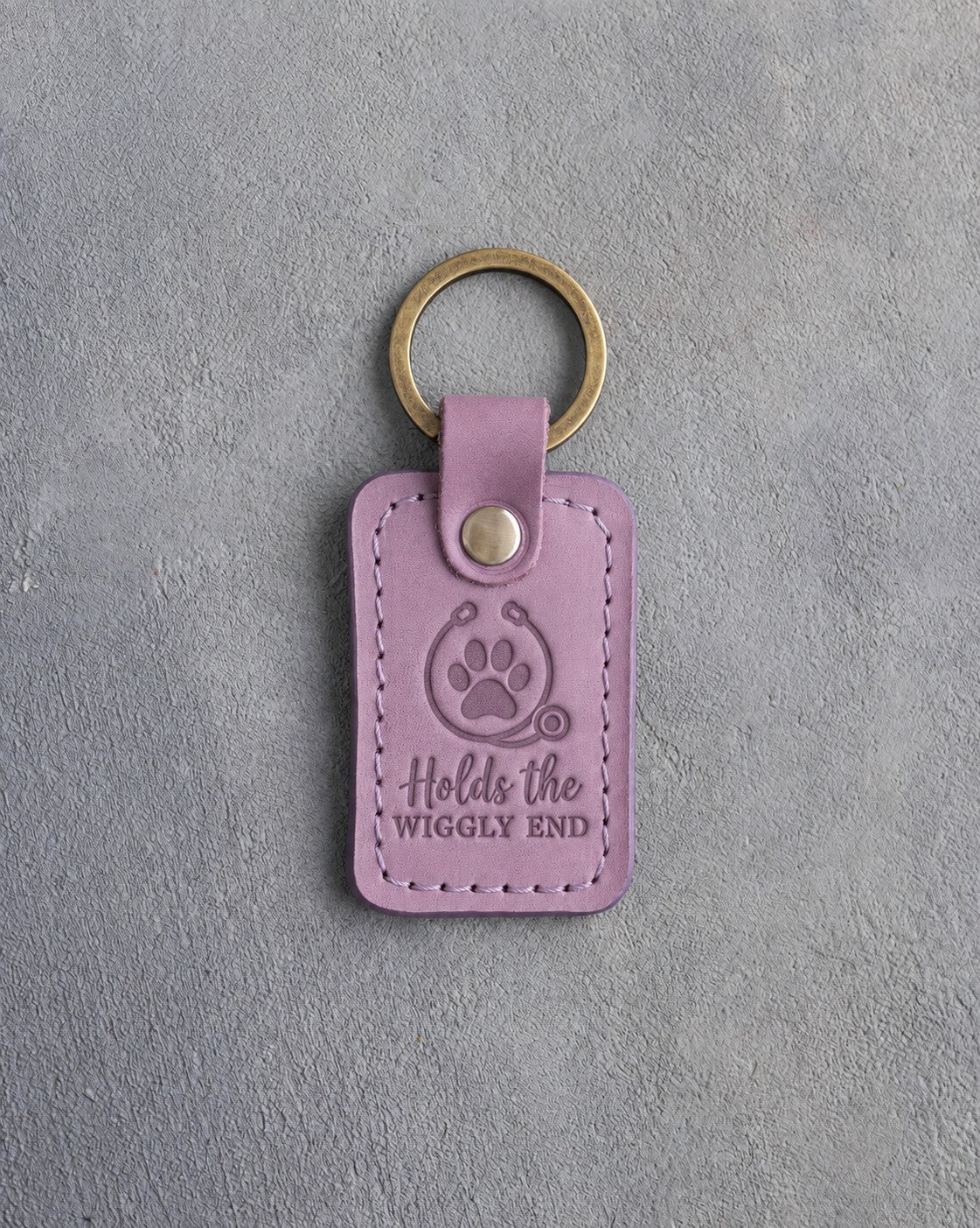 Lilac leather keychain debossed with a paw print circled by a stethoscope and the words Holds the WIGGLY END, on gray concrete