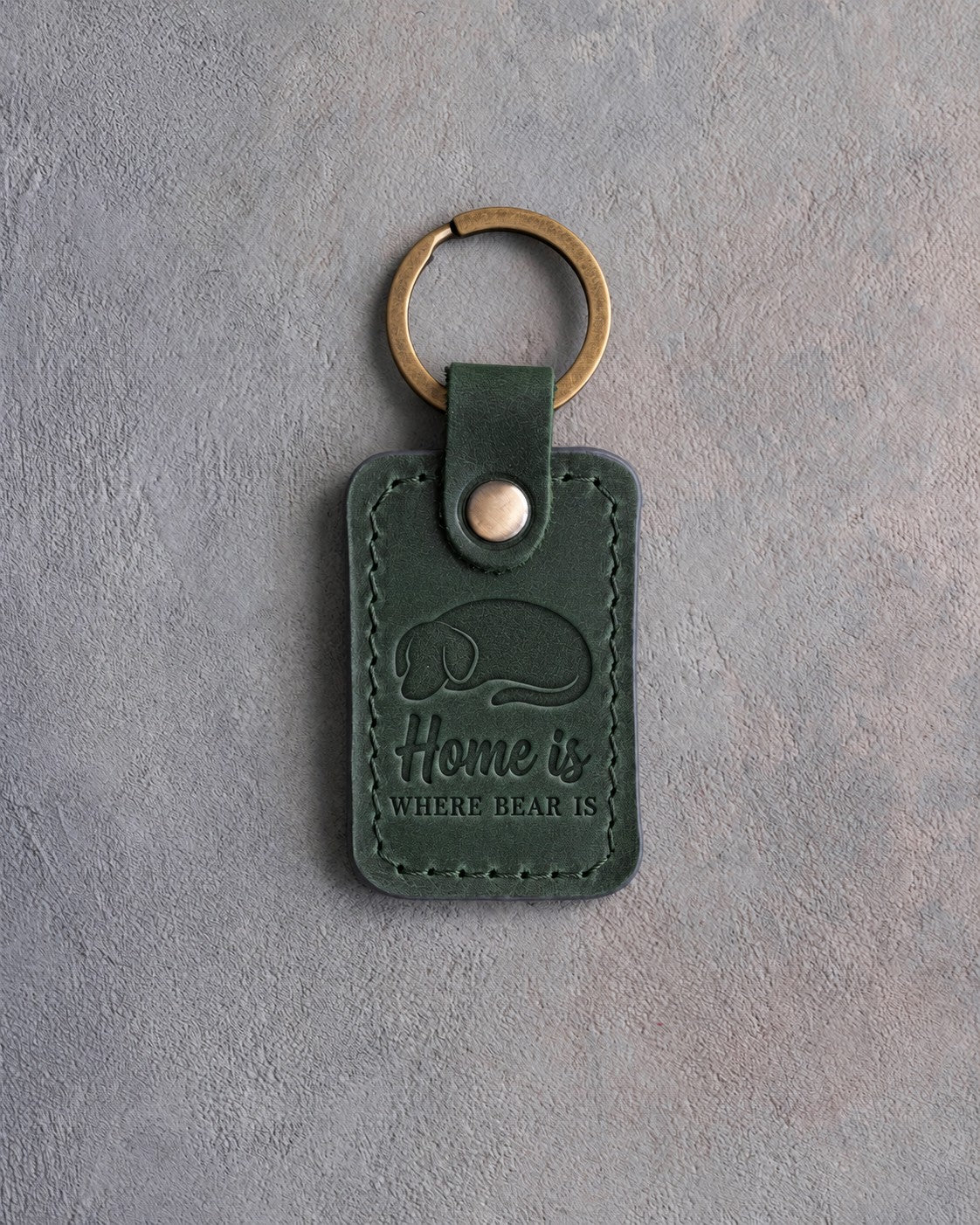 Forest green leather keychain debossed with a curled sleeping dog above Home is and WHERE BEAR IS, on gray concrete