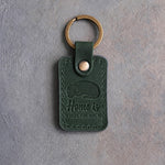 Forest green leather keychain debossed with a curled sleeping dog above Home is and WHERE THE DOG IS, on gray concrete