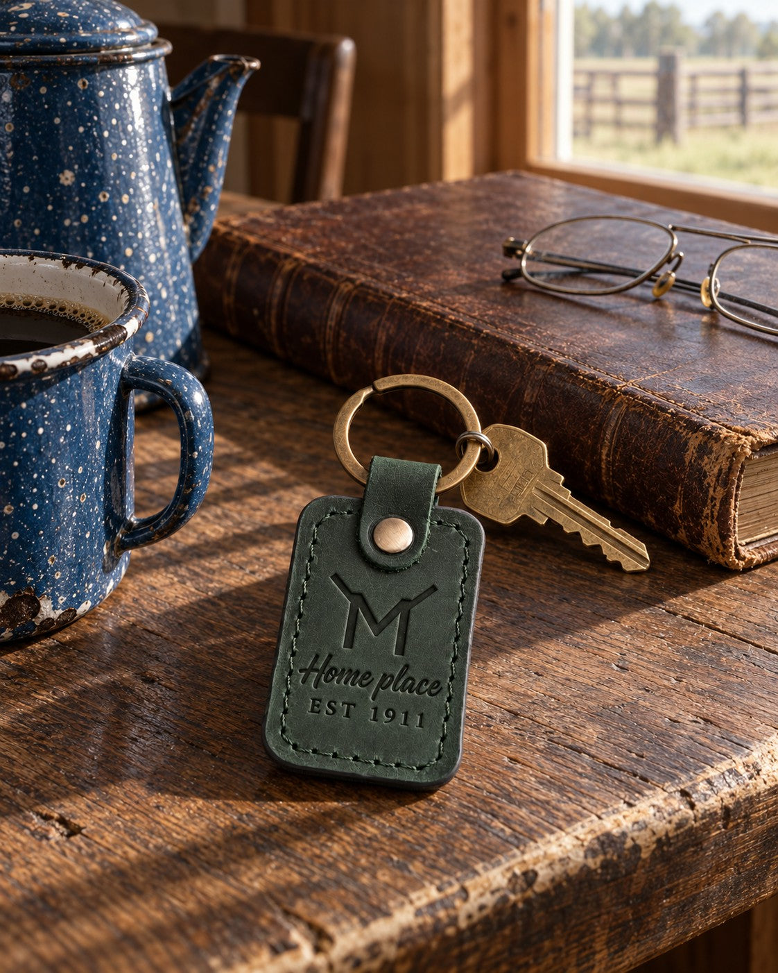 Forest green Home place EST 1911 keychain on a dark oak kitchen table beside a closed leather ledger and reading glasses