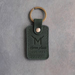Forest green leather keychain debossed with a Flying M ranch brand above Home place and EST 1911, on gray plaster