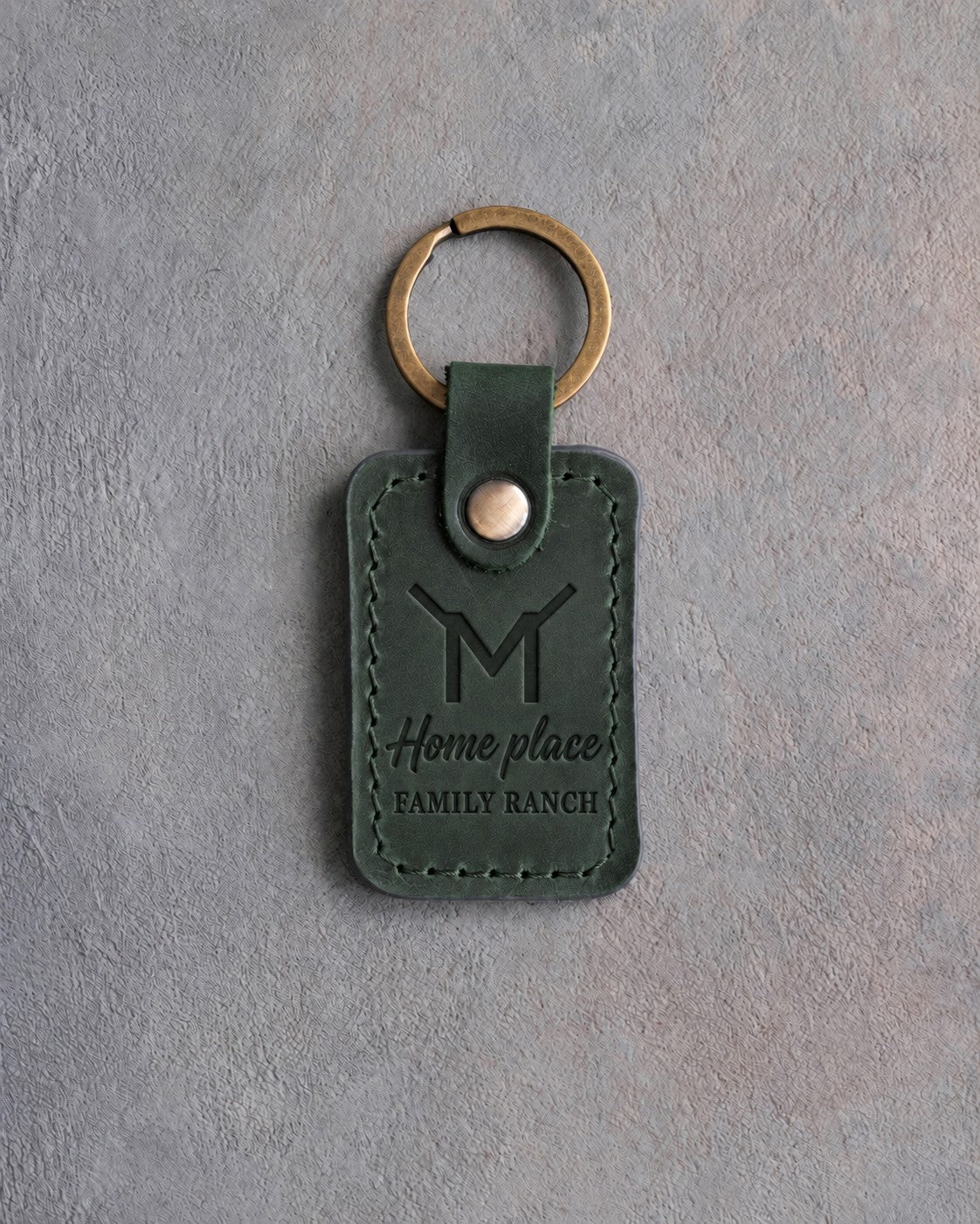 Forest green leather keychain debossed with a Flying M ranch brand above Home place and FAMILY RANCH, on gray concrete