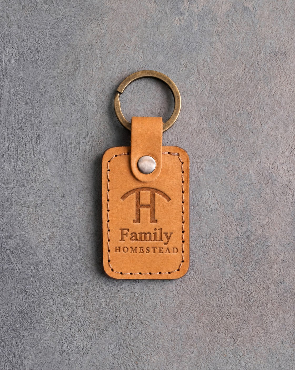Indian Summer tan leather keychain debossed with a Swinging H ranch brand above Family and HOMESTEAD, on gray concrete