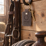 Country gray Horse DAD keychain hanging from a forged bridle hook on rough pine boards in a tack room, a saddle soft on its rack below
