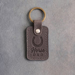 Country gray leather keychain debossed with a heels-up horseshoe above Horse and DAD, on gray plaster