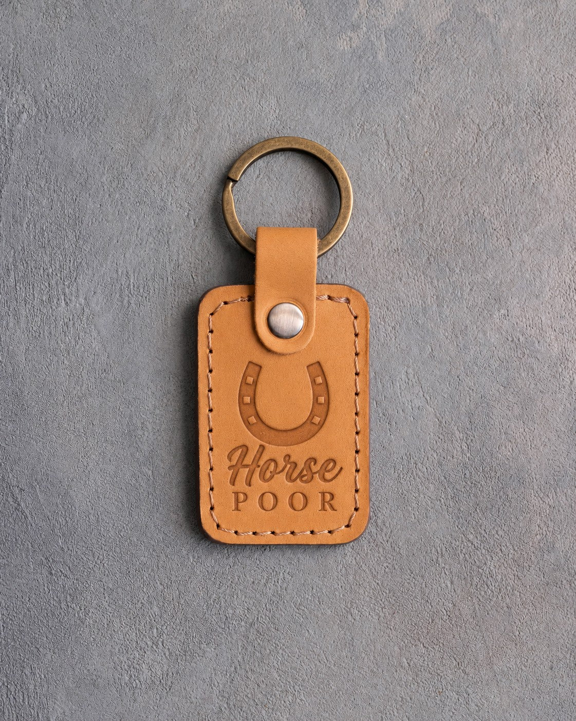 Indian Summer tan leather keychain debossed with a horseshoe above Horse and POOR, on gray plaster