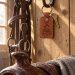 Tate HORSES keychain in Arizona leather hanging from a forged iron bridle hook on a pine tack room wall beside a worn headstall