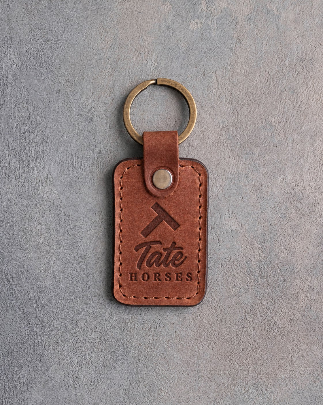 Arizona tan leather keychain debossed with a Tumbling T ranch brand above the name Tate and HORSES, on gray plaster