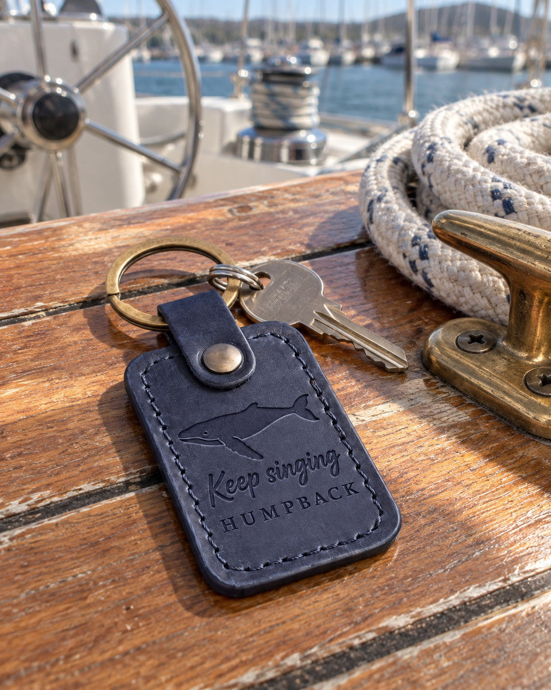 Blueberry Keep singing HUMPBACK keychain in full-grain leather on the worn teak cockpit table of a sailboat beside a coiled white dock line