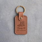 Caramel leather keychain debossed with a pair of service boots above the words Hung up MY BOOTS, on gray concrete