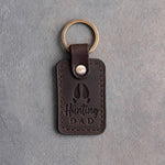 Dark espresso leather keychain debossed with a whitetail deer track above the words Hunting DAD, on gray plaster
