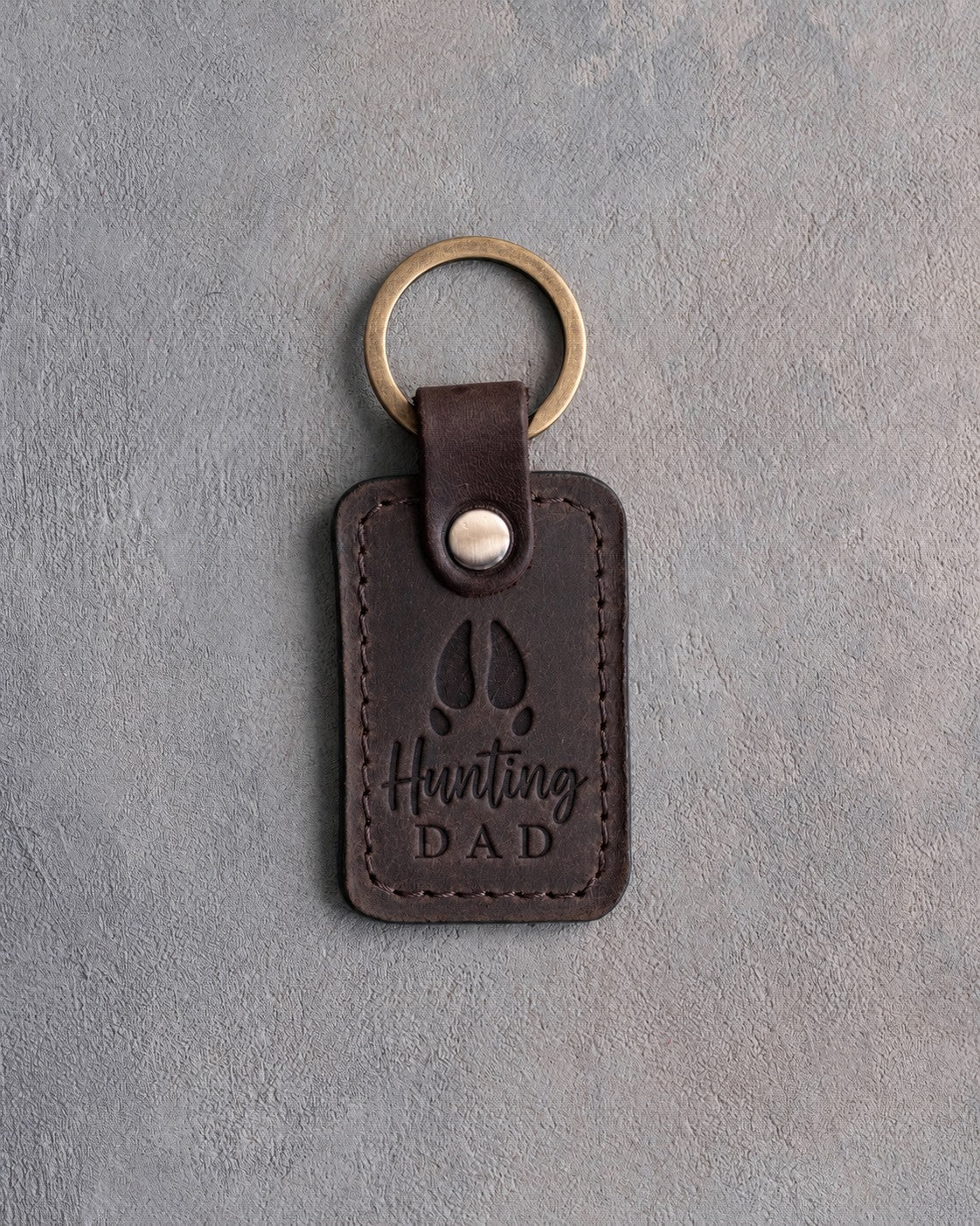 Dark espresso leather keychain debossed with a whitetail deer track above the words Hunting DAD, on gray plaster