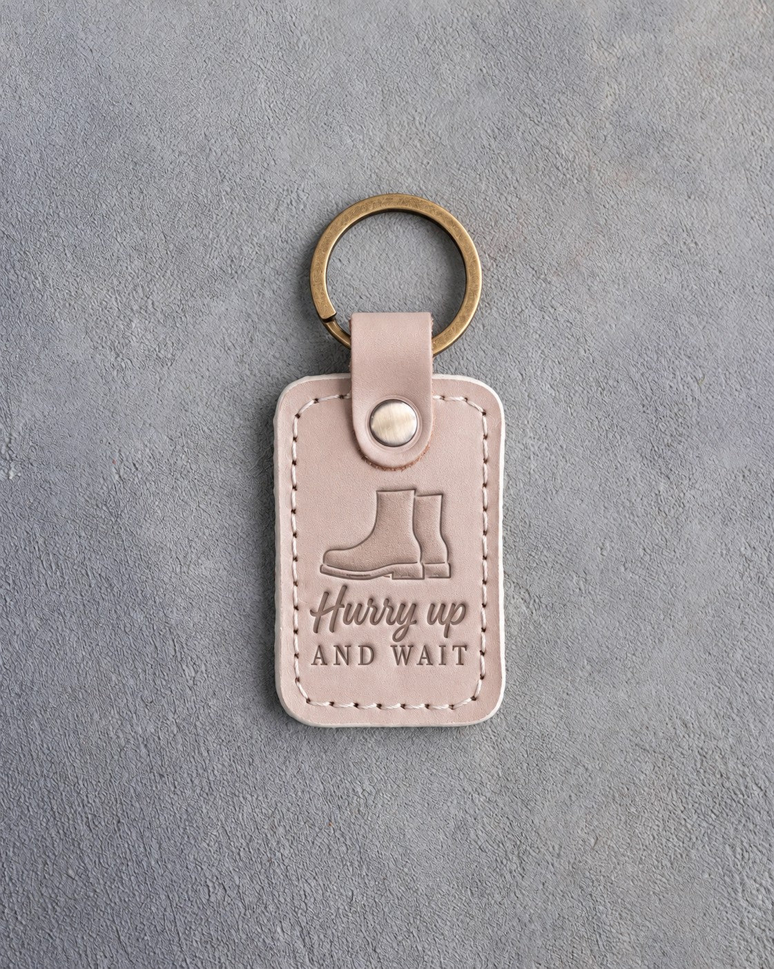 Gray sand leather keychain debossed with a pair of service boots above the words Hurry up AND WAIT, on gray concrete