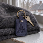 Navy Ignores Me Lovingly keychain on a windowsill with a cat's tail just in frame