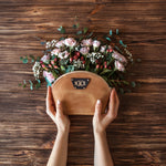 Caramel round leather makeup pouch with a monogram patch, held above a floral arrangement