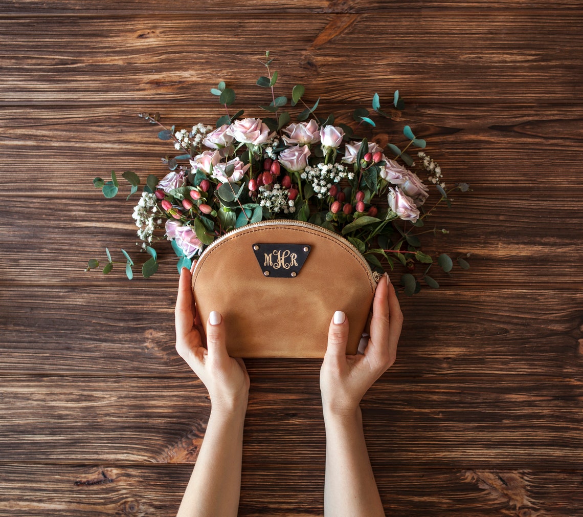 Caramel round leather makeup pouch with a monogram patch, held above a floral arrangement