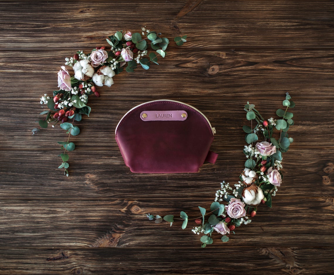 Personalized round leather makeup bag in sangria with a gold nameplate, set in a rose and eucalyptus wreath