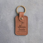 Caramel leather keychain debossed with a golf ball on the lip of the cup under a pin flag above Inside THE LEATHER, on gray plaster