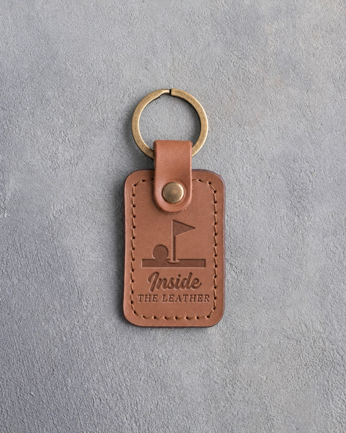 Caramel leather keychain debossed with a golf ball on the lip of the cup under a pin flag above Inside THE LEATHER, on gray plaster