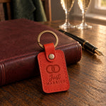 Just MARRIED keychain in bright red leather on a dark oak table beside a closed leather register and a fountain pen