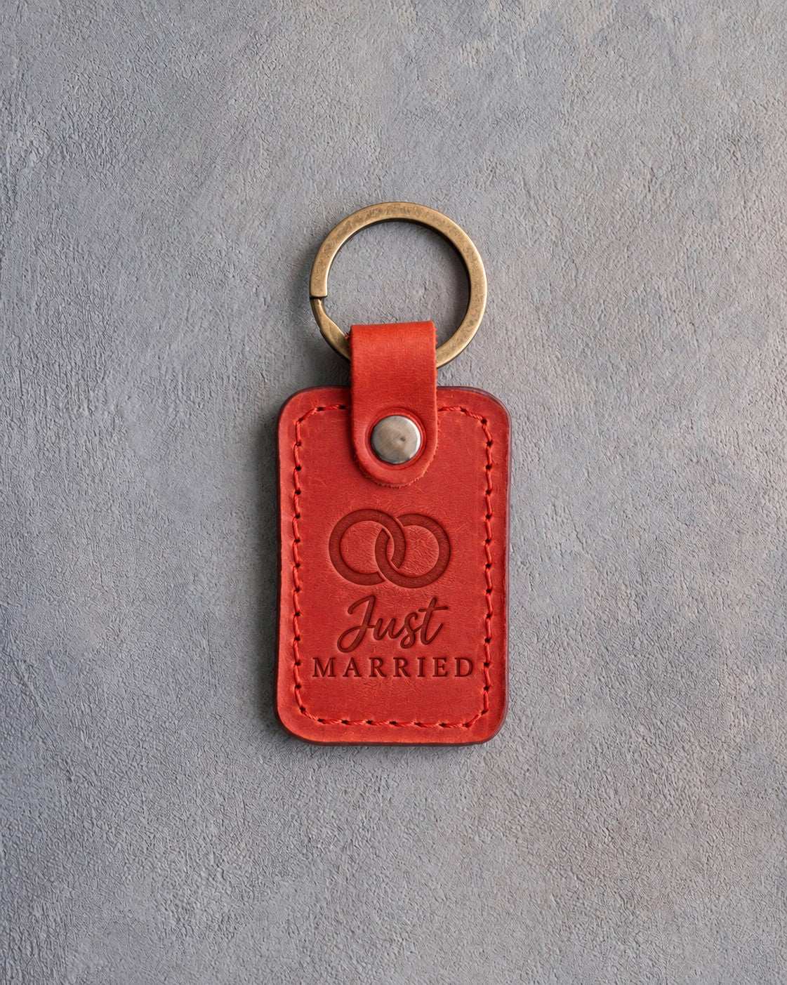 Poppy red leather keychain debossed with two interlocked wedding rings above the words Just MARRIED, on gray concrete