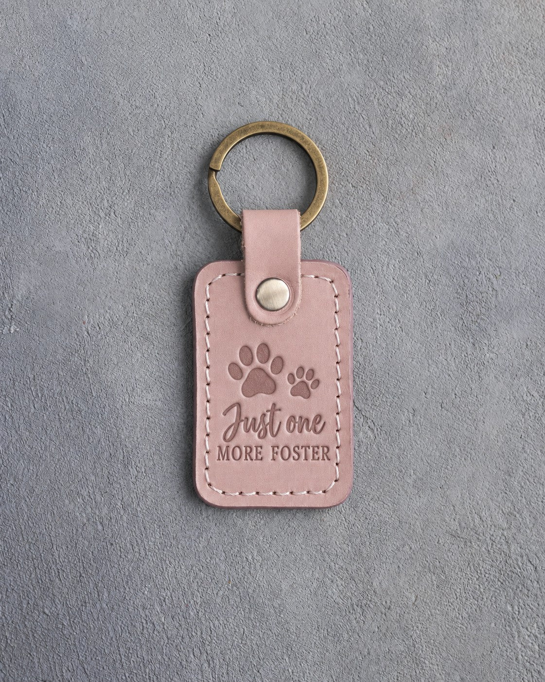 Blush pink leather keychain debossed with a pair of paw prints and the words Just one MORE FOSTER, on gray concrete