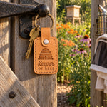 Golden tan Keeper of Bees keychain beside a jar of honey and a hive tool on a wooden bench