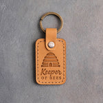 Indian Summer tan leather keychain debossed with a domed skep beehive above Keeper OF BEES, on gray concrete