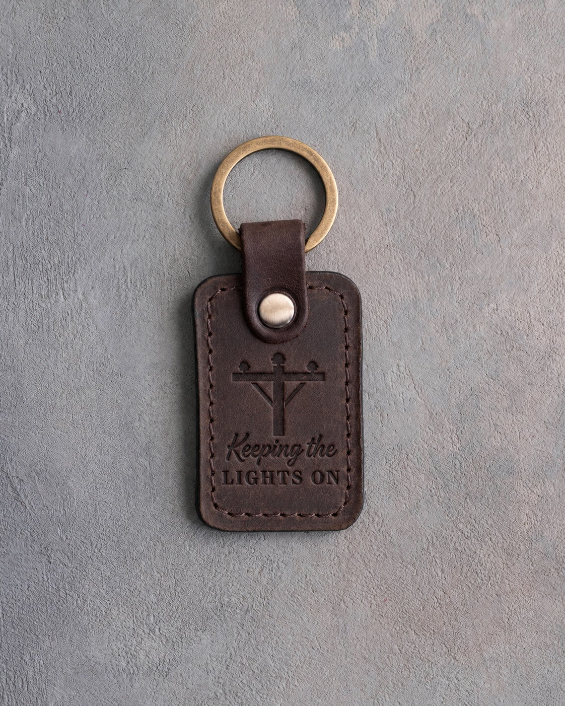 Dark espresso leather keychain debossed with a utility pole above the words Keeping the and LIGHTS ON, on gray plaster