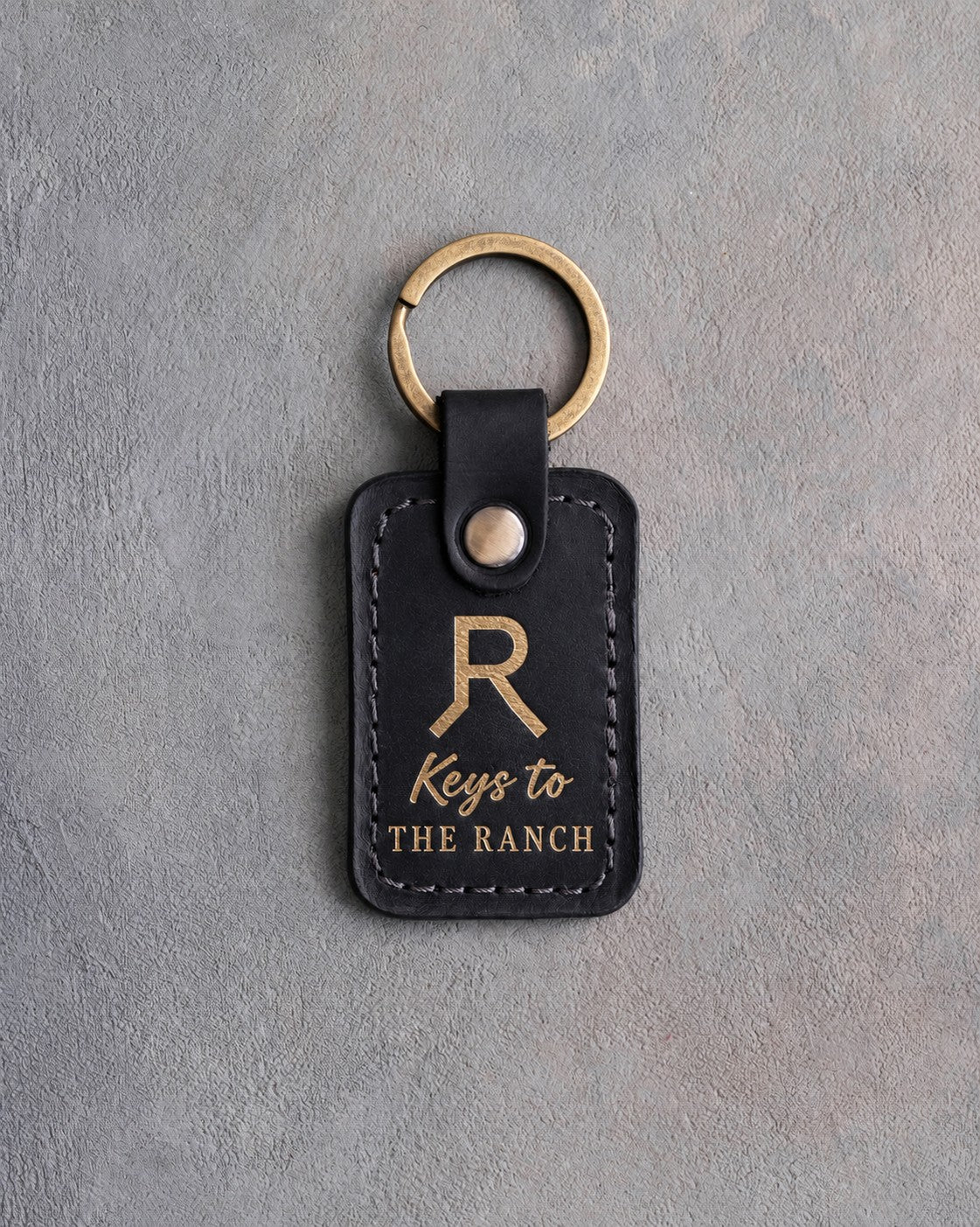 Black leather keychain with a gold foil Walking R ranch brand above Keys to and THE RANCH, on gray plaster