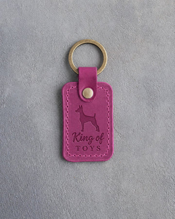 Fuchsia leather keychain debossed with a miniature pinscher and the words King of TOYS, on gray concrete