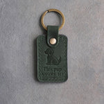 Forest green leather keychain debossed with a sitting puppy above This pup and KNOWS SIT, on gray concrete