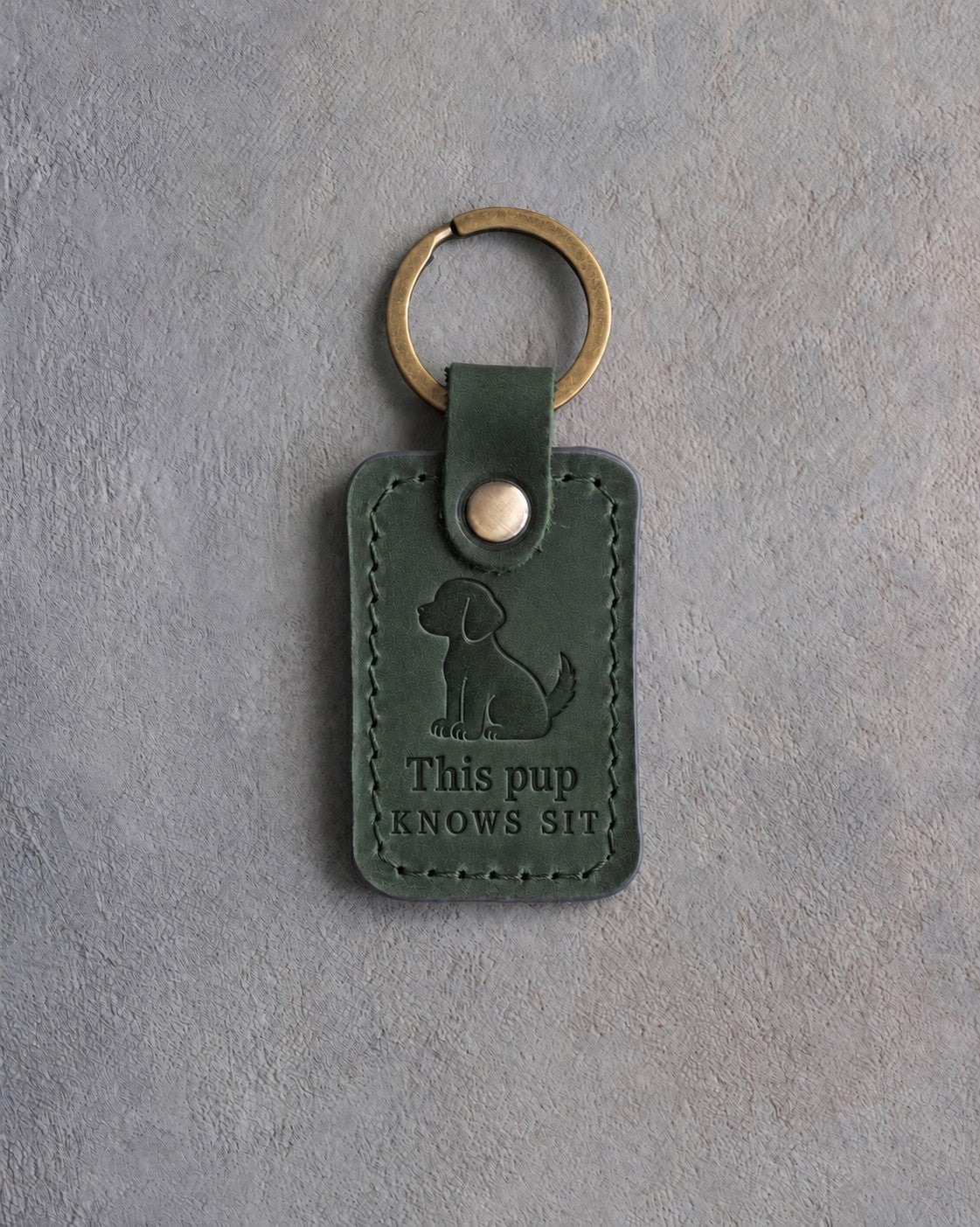Forest green leather keychain debossed with a sitting puppy above This pup and KNOWS SIT, on gray concrete