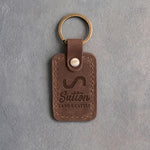Sicilian brown leather keychain debossed with a Lazy S ranch brand above the name Sutton and LAND & CATTLE, on gray plaster