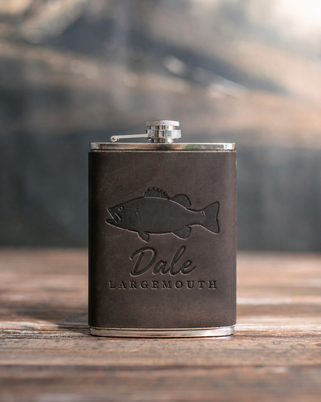 Dark espresso leather-wrapped hip flask debossed with a largemouth bass above Dale and LARGEMOUTH, on a wooden table